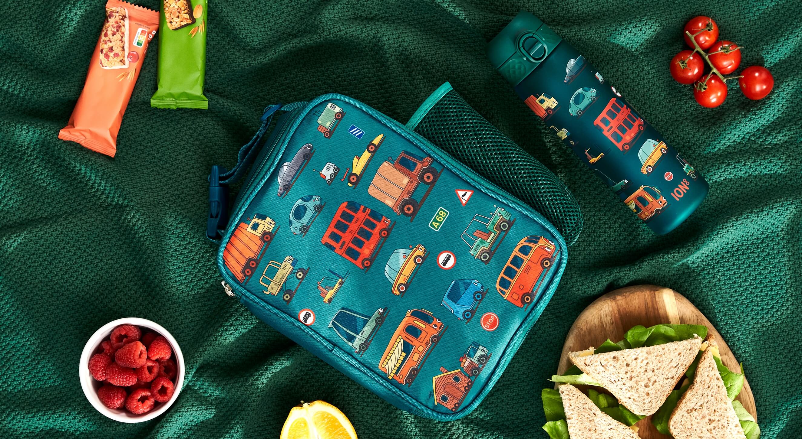 A teal lunch bag, adorned with vehicle illustrations, rests on a green fabric. Nearby are food items and a matching water bottle labeled