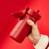A red cylindrical water bottle with a flip-top lid is held against a solid red background.
