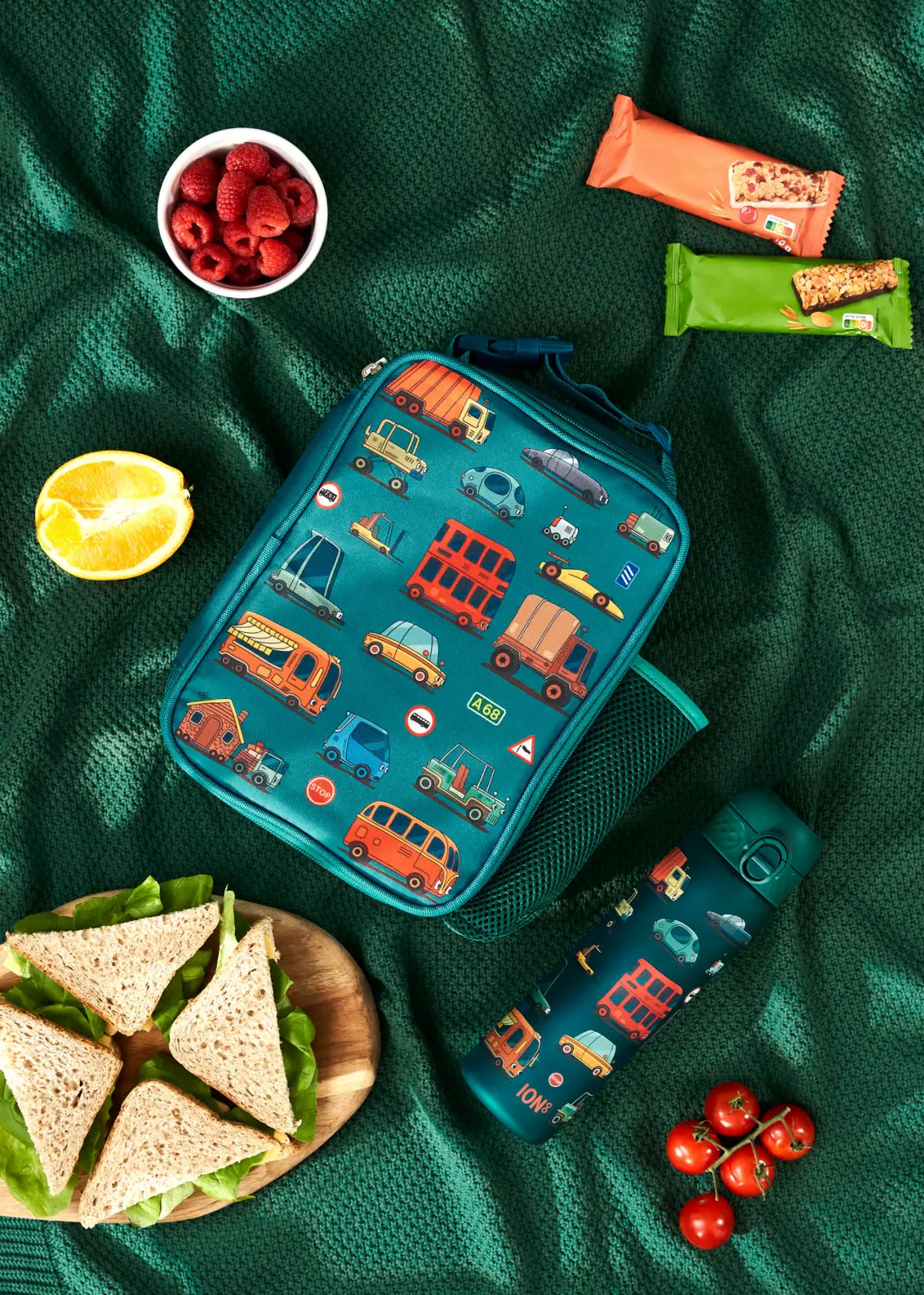 A teal lunch bag, decorated with various vehicles, rests on a green blanket alongside a matching water bottle, sandwiches, fruit, and granola bars.