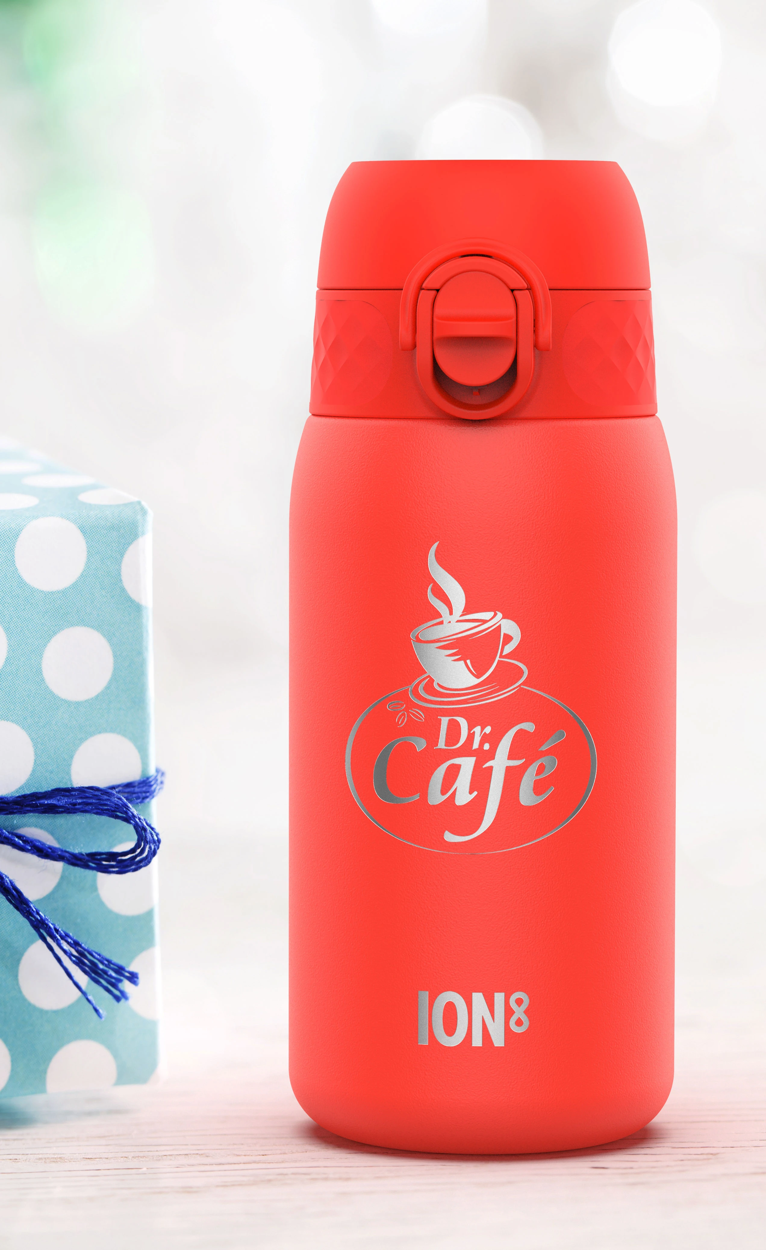 A red ION8 thermos sits on a white wooden surface near a wrapped gift. It displays a silver logo: "Dr. Café" with a coffee cup image.