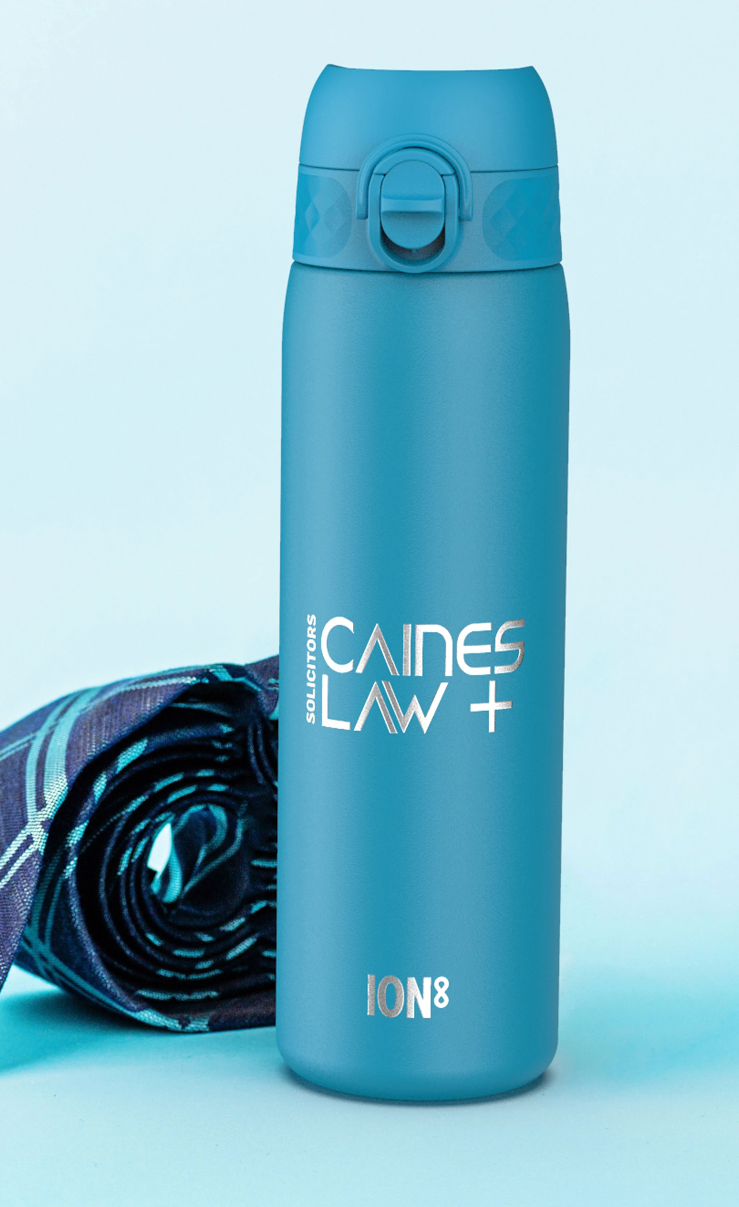 A teal water bottle stands on a light blue surface next to a rolled-up tie. The bottle displays “CAINES LAW + SOLICITORS” and “ION8”.