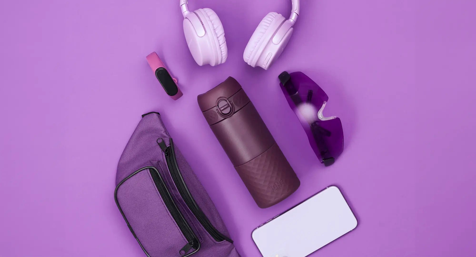 Various purple items rest on a purple background. A water bottle sits among a fanny pack, headphones, fitness tracker, sunglasses, and a phone.