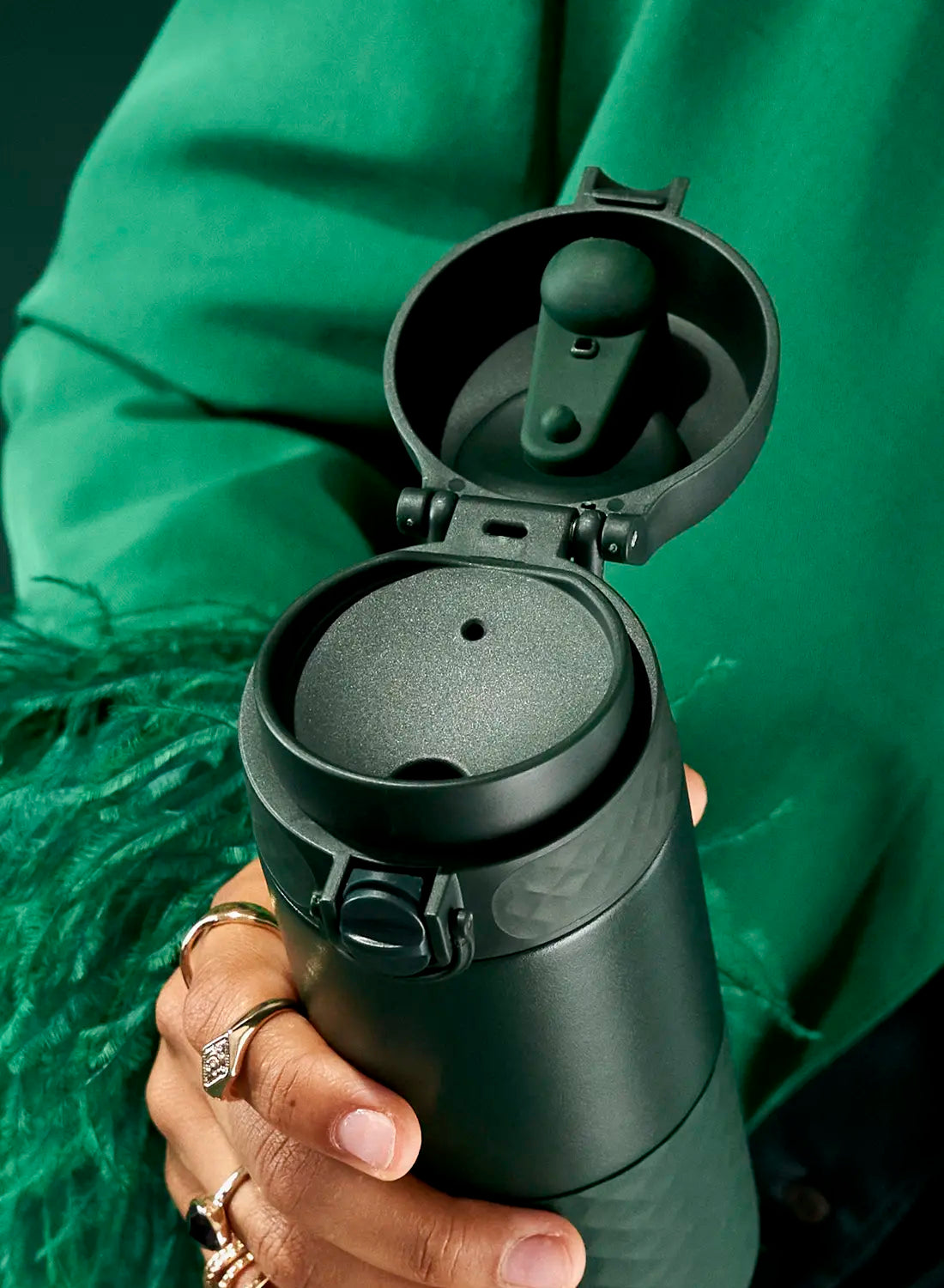 A dark-green travel mug, open, is held in a hand. The hand is adorned with rings. The background is a green garment with a feather boa.