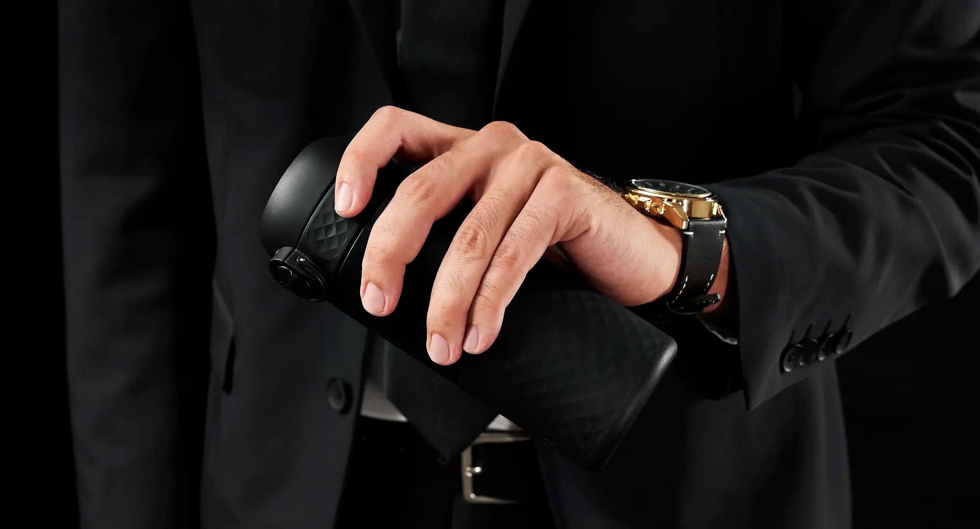 A hand holds a black, textured travel mug. The person is wearing a black suit and a gold watch. The setting appears to be a dark, possibly indoor space.