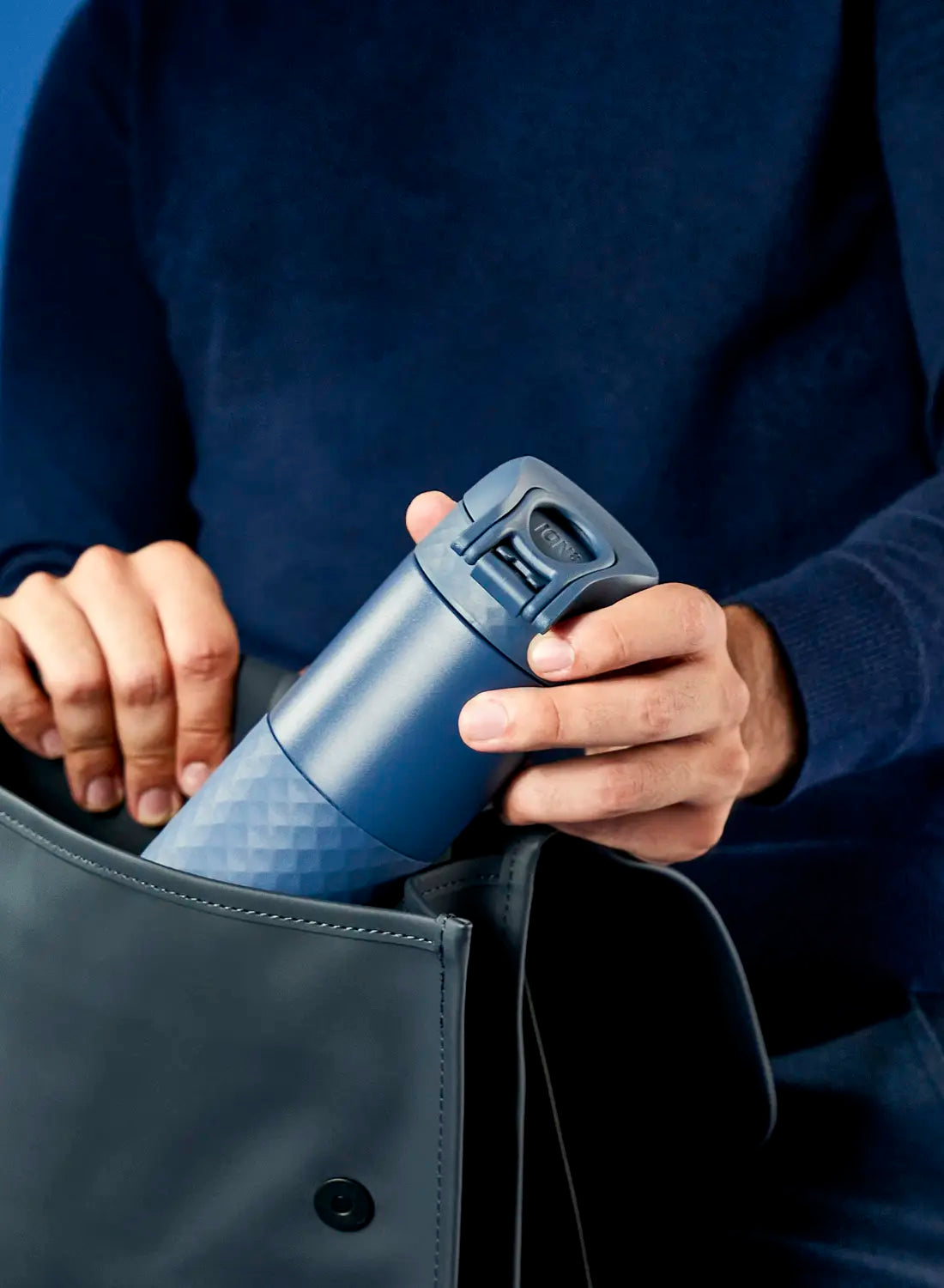 A blue, textured thermos is being placed into a dark gray bag by a person wearing a dark blue sweater.