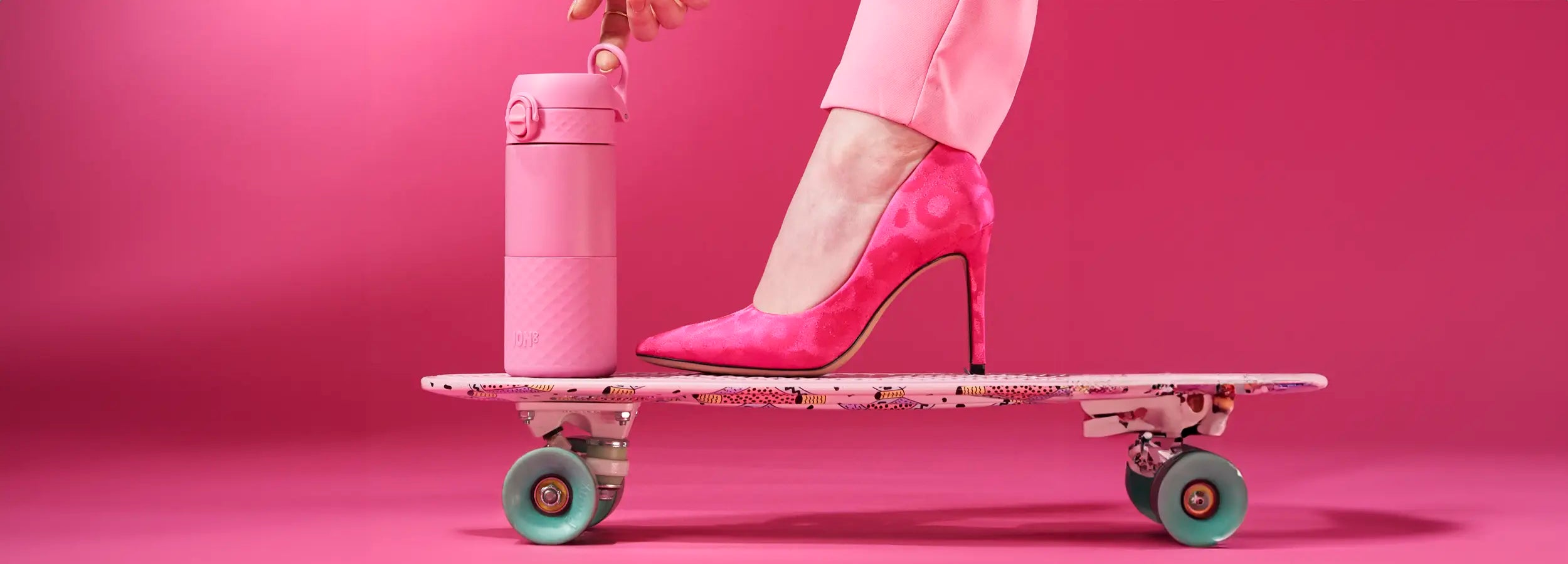 A pink water bottle is being placed on a patterned skateboard, which is also supporting a woman's pink high heel. The scene is set against a solid pink background. The bottle has the word "JOM8" on it.