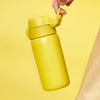 A yellow insulated water bottle is being held by a hand against a yellow background.