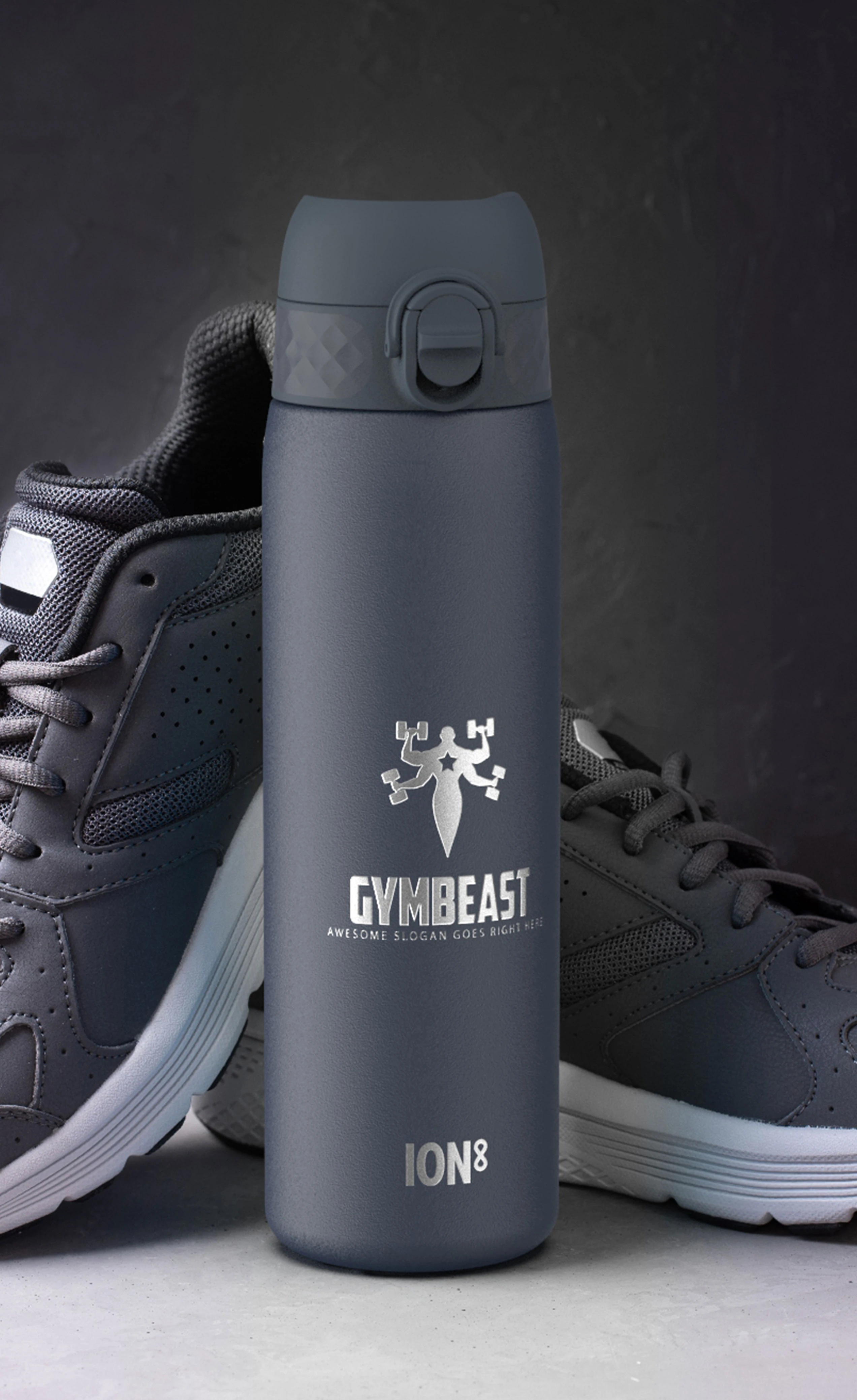 A dark-grey water bottle stands between two matching athletic shoes on a grey surface. The bottle features a logo: "GYMBEAST" and "AWESOME SLOGAN GOES RIGHT HERE," plus "ION8."
